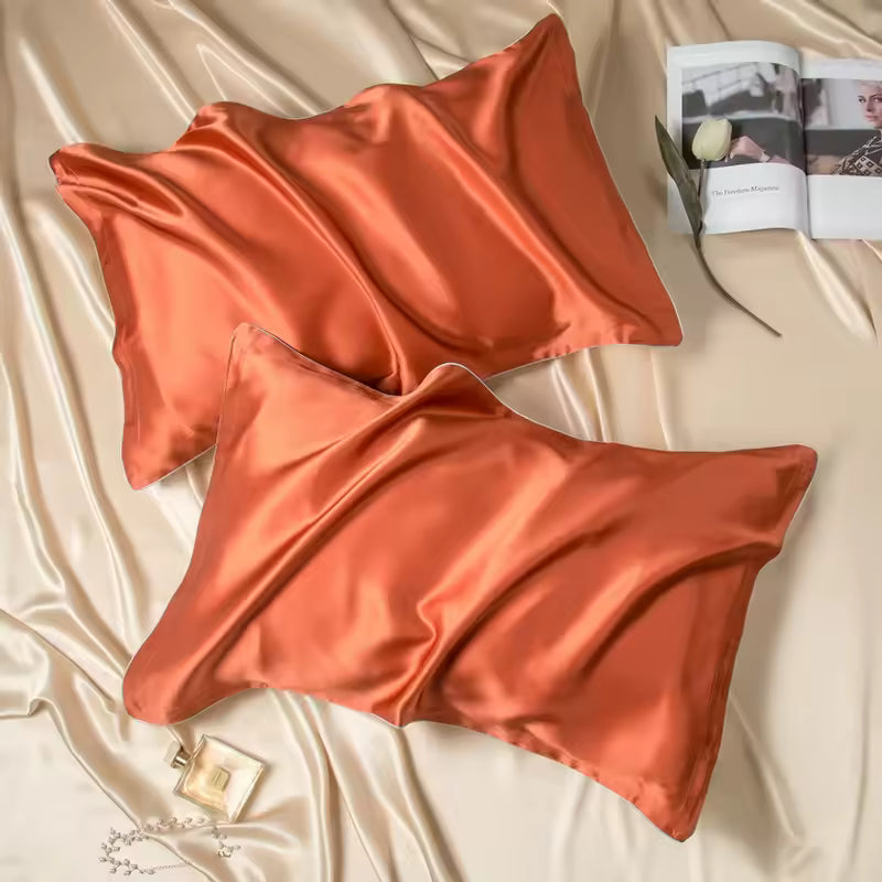 Two rust orange silk pillowcases on champagne silk fabric with an open magazine and perfume bottle.
