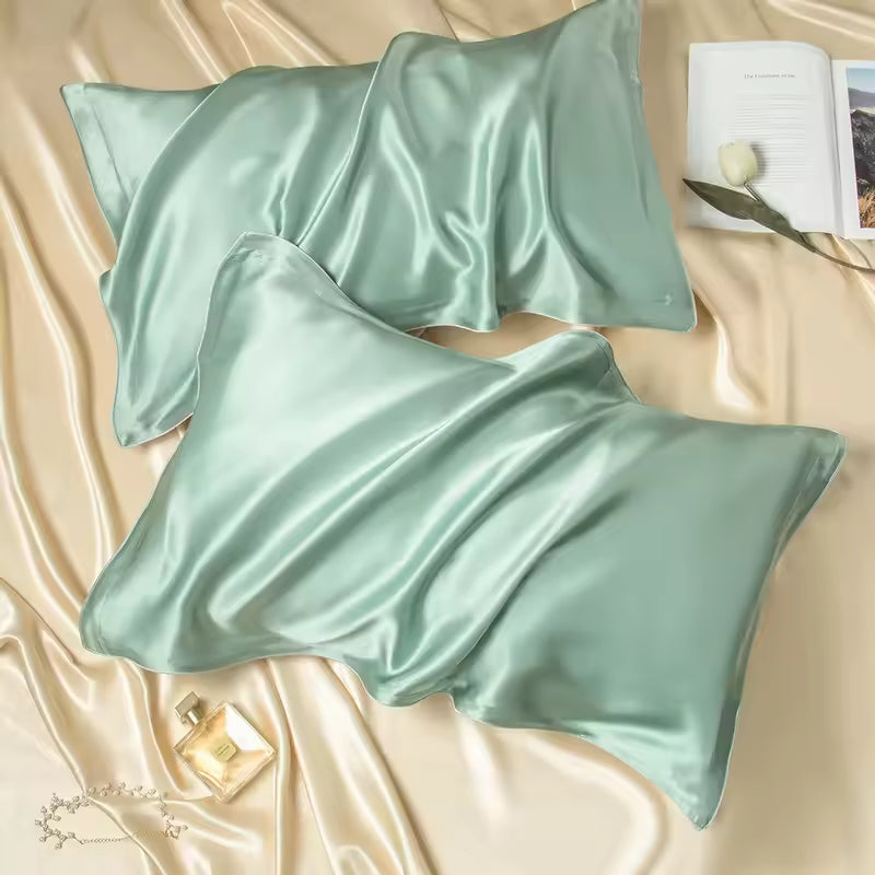 Two sage green silk pillowcases arranged on beige silk fabric alongside a book and perfume bottle.