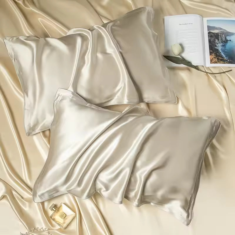 Two champagne silk pillowcases lying on matching draped silk with an open book and perfume.