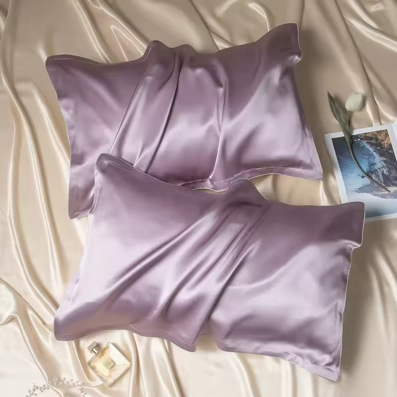 Two lilac purple silk pillowcases resting on champagne silk fabric with a picture card and perfume.