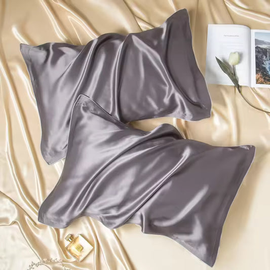 Two metallic gray silk pillowcases laid on silky champagne fabric beside a book and perfume.