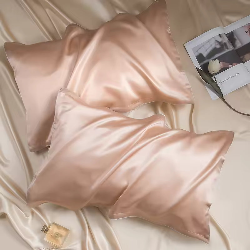 Two rose gold silk pillowcases lying on champagne silk fabric with an open magazine and perfume.
