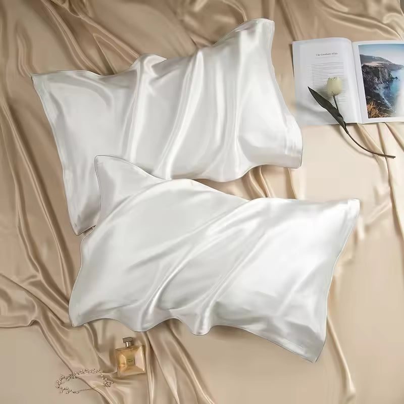 Two white silk pillowcases set on champagne silk fabric alongside an open book and perfume bottle.
